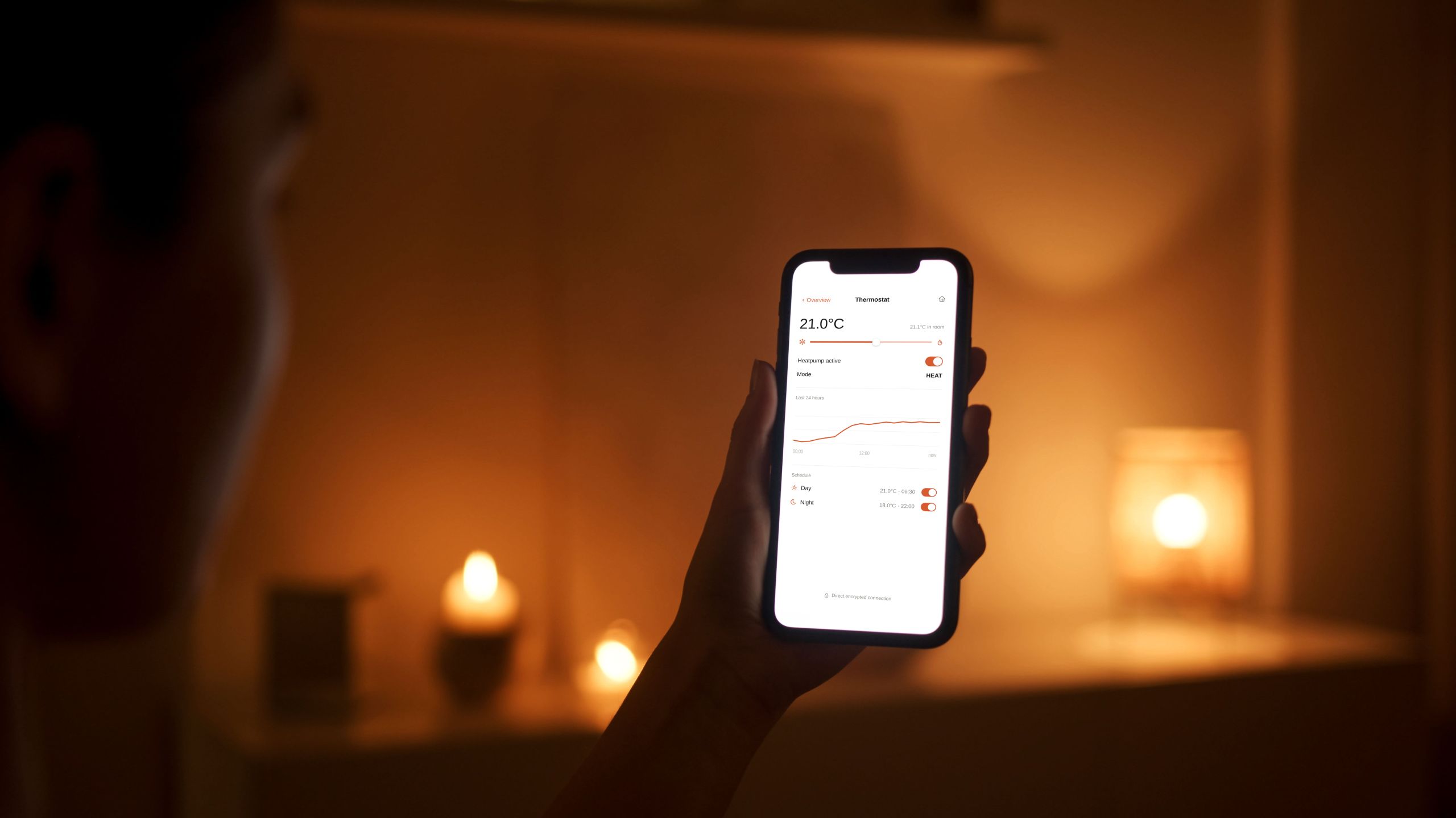 A hand holding a smartphone showing a thermostat app in a dark candle-lit living room