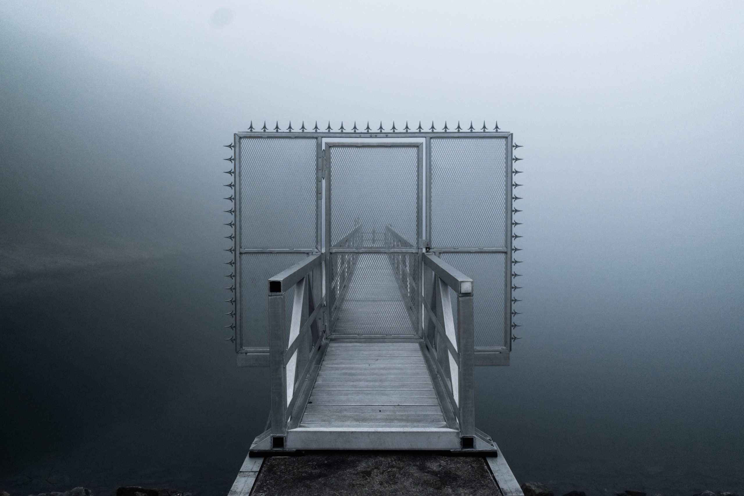 A spiked steel gate shut across a dock walkway, fog and flat water on either side