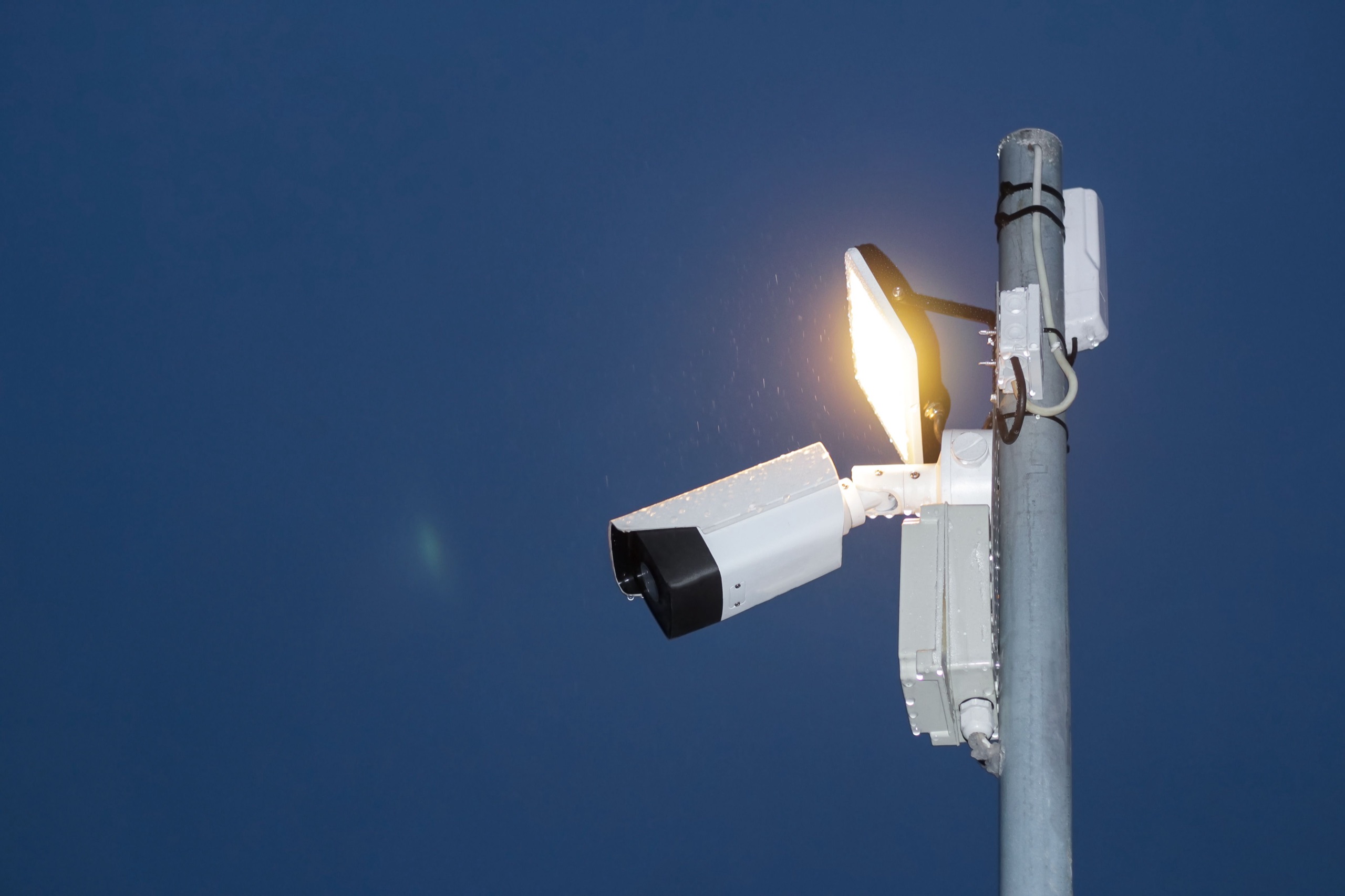An IP security camera and floodlight on a pole against a deep blue dusk sky